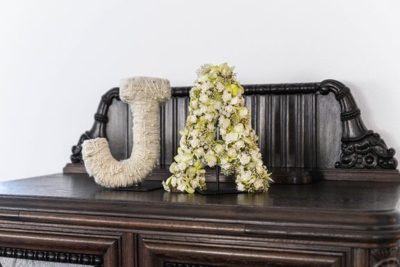 Die Buchstaben "J" und "A", dekoriert mit weißen und grünen Blumen, stehen auf einem Holztisch.