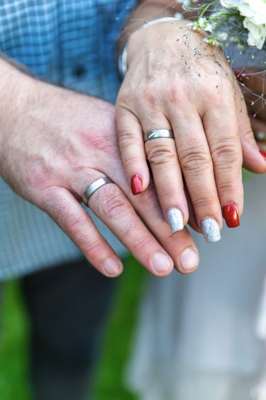 Hände eines Paares mit Eheringen, eine Hand hat rote und die andere silberne Nägel.